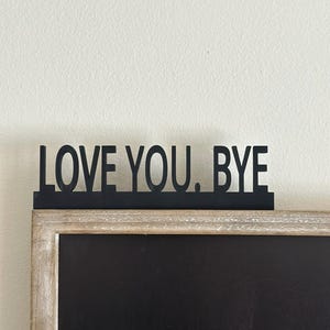 May include: Black metal sign with the words "LOVE YOU. BYE" above a framed chalkboard. The frame is a light wood color, and the chalkboard has a black surface. The sign is mounted on top of the frame.