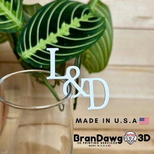 May include: White wooden letter L and D with an ampersand symbol in between, attached to a clear glass stem. The letters are on a wooden surface with a green plant in the background. The text "Made in U.S.A." and "BranDawg 3D Printing Services Made in U.S.A." is visible below the letters.