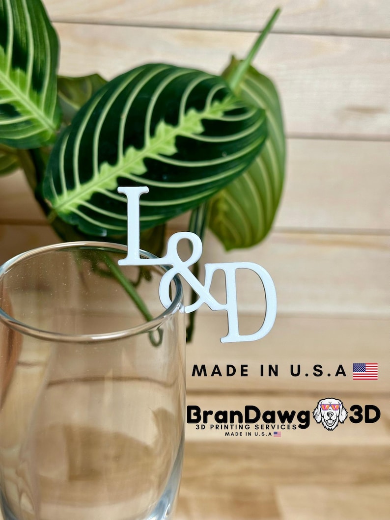 May include: White wooden letter L and D with an ampersand symbol in between, attached to a clear glass stem. The letters are on a wooden surface with a green plant in the background. The text "Made in U.S.A." and "BranDawg 3D Printing Services Made in U.S.A." is visible below the letters.