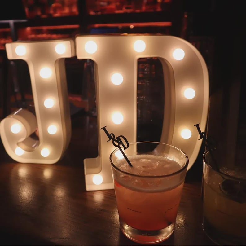 May include: Two large, white, illuminated marquee letters J and D stand behind two glass tumblers filled with cocktails. Each drink contains a black stirring stick featuring the letters J and D at the top, positioned against the glowing lights.