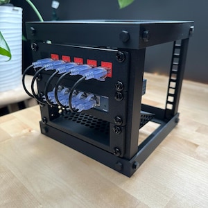 May include: A black server rack with visible network cables and ports. The rack is rectangular with a bolted frame. The network cables are blue and connected to red ports. The rack sits on a light-colored wooden surface.