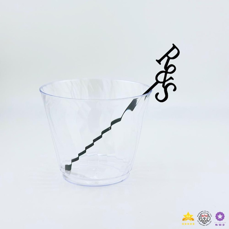 May include: A clear, transparent plastic cup with a faceted, textured surface. Inside the cup is a black, thin, decorative stirring stick featuring a zigzag stem and the letters R&S in a stylised, connected script font at the top end.