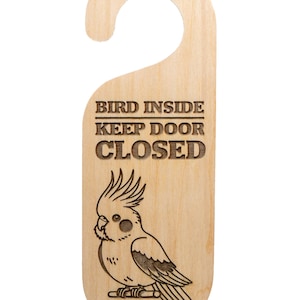 May include: Wooden door hanger with a cutout for the doorknob. The text "BIRD INSIDE KEEP DOOR CLOSED" is engraved above a detailed illustration of a cockatiel. The wood has a light, natural finish.
