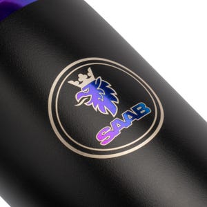 May include: Black cylindrical object with a metallic iridescent logo. The logo features a crown, a bird, and the word "SAAB" in a gradient of purple, blue, and pink. The object has a textured surface.