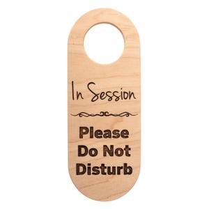 May include: A wooden door hanger with a hole at the top. The text "In Session" is above a decorative line, followed by "Please Do Not Disturb" in a bold, sans-serif font. The wood has a light, natural finish.