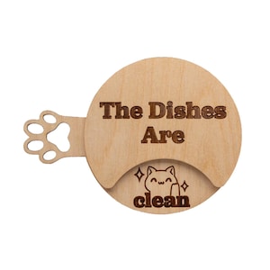 May include: A circular light-colored wooden sign with a paw print cutout on the left side. The text "The Dishes Are" is engraved in the center, and below it, a small engraved cat illustration with the word "clean" and two star symbols.