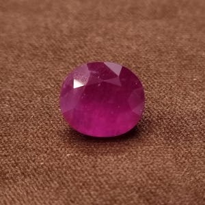 Op de afbeelding: Een gefacetteerde, ronde robijn edelsteen. De edelsteen heeft een diepe, roze-rode kleur, met meerdere facetten die het licht vangen. De edelsteen ligt tegen een bruine achtergrond.