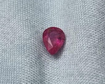 Puede incluir: Una gema de rubí facetada en forma de pera. El rubí es de un color rojo intenso y rico con inclusiones internas visibles. La gema está sobre un fondo gris claro texturizado.