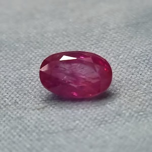 May include: A faceted, oval-shaped ruby gemstone. The ruby is a deep, saturated red color with internal inclusions. The gemstone is displayed on a gray, textured surface, showcasing its brilliance and clarity.