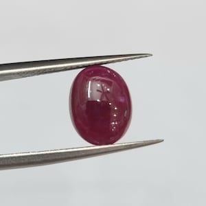 Könnte beinhalten: Ein polierter, ovalförmiger Rubin-Edelstein, der von einer Pinzette gehalten wird. Der Rubin hat eine tiefe, satte rote Farbe mit einer glatten, reflektierenden Oberfläche. Der Edelstein ist ein Naturstein.