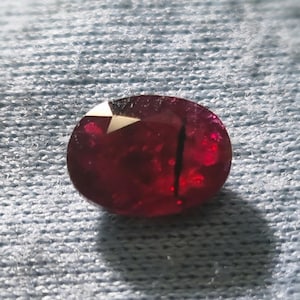 Puede incluir: Una gema de rubí facetada, de forma ovalada. La gema es de un color rojo intenso y saturado con una ligera inclusión interna. El corte de la piedra revela múltiples facetas, que reflejan la luz. El rubí está sobre un fondo azul claro texturizado.