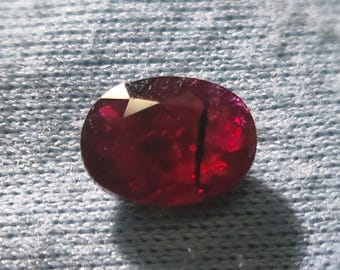 Puede incluir: Una gema de rubí facetada, de forma ovalada. La gema es de un color rojo intenso y saturado con una ligera inclusión interna. El corte de la piedra revela múltiples facetas, que reflejan la luz. El rubí está sobre un fondo azul claro texturizado.