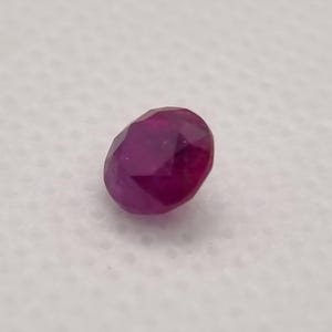 May include: A faceted, oval ruby gemstone. The gem is a deep red colour, with multiple facets that catch the light. The ruby is set against a white background.