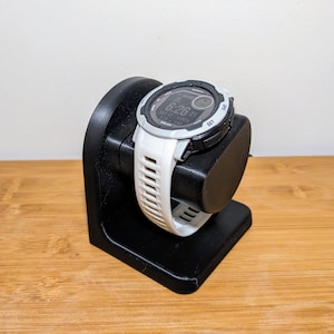 May include: A black watch charging stand with a white and black smartwatch. The watch displays the time as 6:26. The watch has a white band and a black bezel. The stand is made of black plastic and has a curved design.