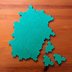 May include: A green puzzle shaped like a map, with many interlocking pieces. Some pieces are separated from the main structure, resting on a wooden surface. The puzzle's color is a vibrant teal, contrasting with the warm wood tones.