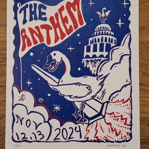 May include: A blue and red linocut print titled "The Anthem." The print features a flag with the title, a goose holding a sword, and the date "NOV 12.13 2024." The print also includes the text "21/40" and the artist's signature.