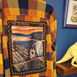 May include: A yellow, orange, and blue plaid flannel shirt with a black and white image of a cat in the style of "The Scream" painting. The image is stitched onto the back of the shirt and has the text "The Scream Cat" above it.
