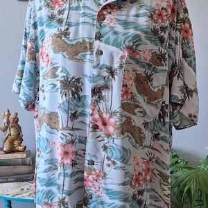 May include: A short-sleeved button-down shirt featuring a light blue background with a repeating pattern of pink hibiscus flowers, brown rocky islands, palm trees, and light blue ocean waves. The shirt includes a single chest pocket on the left side.
