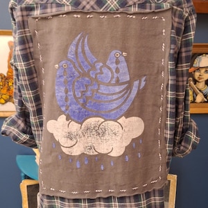 May include: A blue and green plaid flannel shirt with a gray patch sewn on the back. The patch features a blue silhouette of two birds with tear drops on a white cloud with blue raindrops.