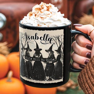 May include: Black ceramic mug with a witch-themed design. The mug features the name "Isabella" and a graphic of witches with the text "Girls Will Be Girls." The mug is filled with a whipped cream topping.