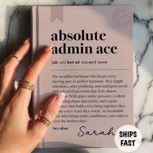 May include: A light pink notebook with the words "absolute admin ace" in black. The definition of "admin ace" is written below the title. The notebook is being held by a hand with gold rings. The text "Ships Fast" is also visible.