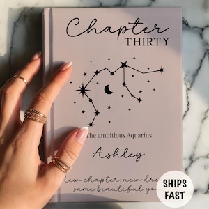 May include: A light mauve hardcover book featuring the black text Chapter THIRTY, a constellation graphic, the phrase the ambitious Aquarius, the name Ashley, and the text new chapter, new dreams, same beautiful you printed on the front cover.