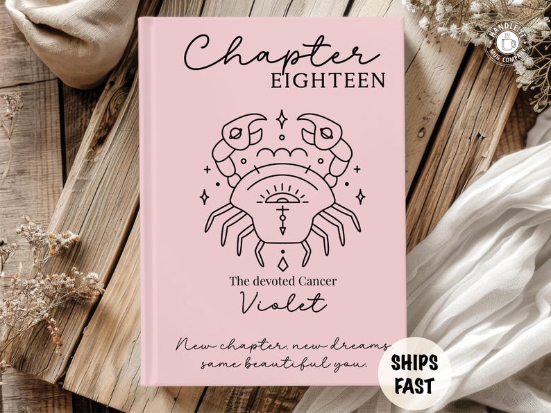 May include: A light pink hardcover book featuring the text Chapter EIGHTEEN at the top, a black line art illustration of a crab zodiac symbol in the center, the text The devoted Cancer Violet below it, and New chapter, new dreams, same beautiful you.