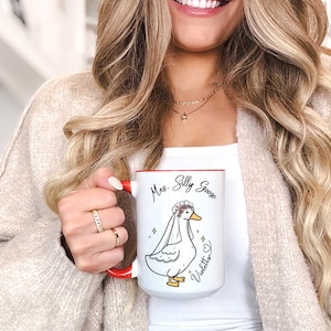 A newly married woman smiling while holding her wedding, engagement or bridal shower gift mug with our Mrs Silly Goose design and her name showing next to it. The mug makes for a great gift for newlyweds, comes as a single or set. It also comes in 11oz, 15oz or 20oz variants and allows for personalization.