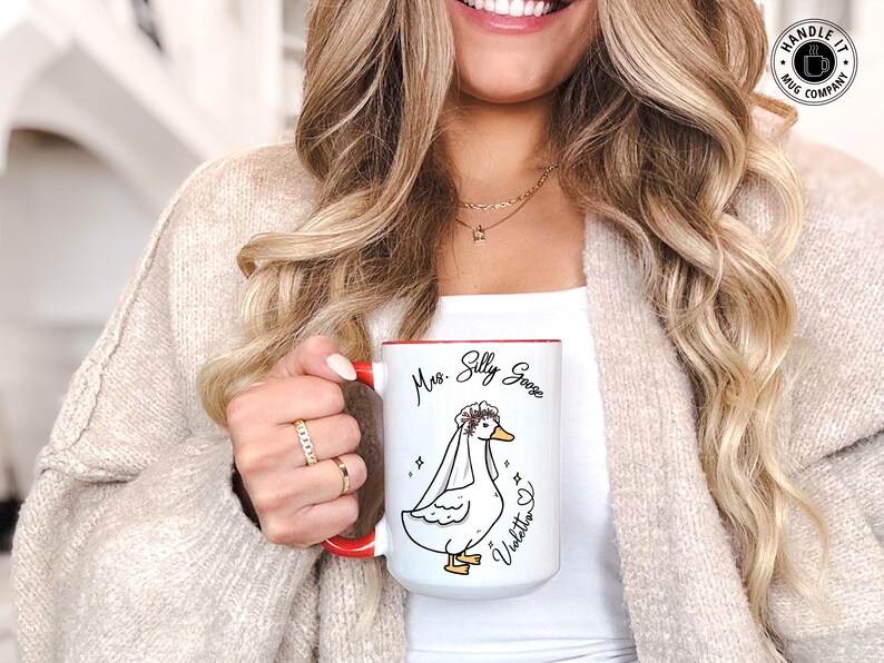 A newly married woman smiling while holding her wedding, engagement or bridal shower gift mug with our Mrs Silly Goose design and her name showing next to it. The mug makes for a great gift for newlyweds, comes as a single or set. It also comes in 11oz, 15oz or 20oz variants and allows for personalization.