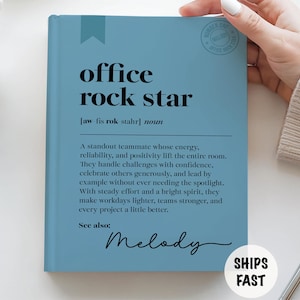 Op de afbeelding: Een lichtblauw notitieboek met de zwarte woorden "office rock star". De definitie van "office rock star" staat onder de titel. Het notitieboek heeft een blauwe lint-bladwijzer en de tekst "SHIPS FAST" in een witte cirkel.
