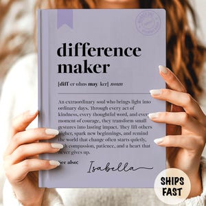 May include: A light purple book with the words "difference maker" in black. The book has a definition and a quote about making a difference. The name "Isabella" is written in cursive. The book is held by a person.