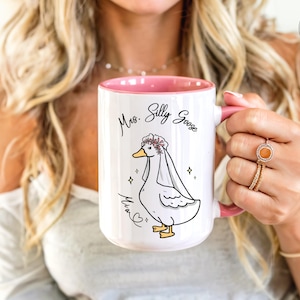 A woman proudly holding her Mrs Silly Goose mug gift. It shows are pink accent mug in 15oz. It appears that she got it as a gift for her engagement, wedding or bridal shower. It also allows for personalization.