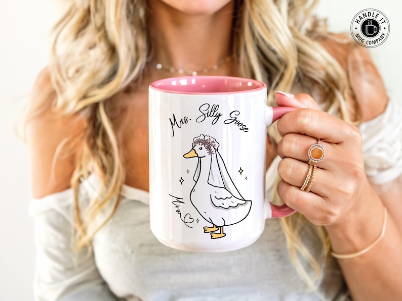 A woman proudly holding her Mrs Silly Goose mug gift. It shows are pink accent mug in 15oz. It appears that she got it as a gift for her engagement, wedding or bridal shower. It also allows for personalization.