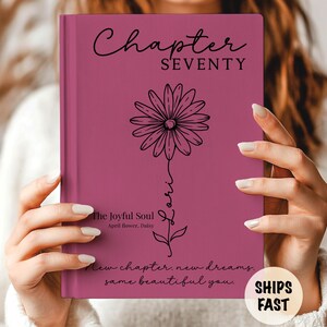 May include: A dark pink hardcover journal featuring a black line art illustration of a daisy flower. The text reads Chapter SEVENTY, Lori, The Joyful Soul, April flower, Daisy, and New chapter, new dreams, same beautiful you in black script.
