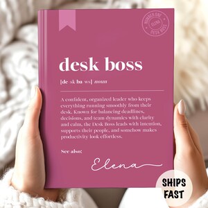 以下が含まれることがあります： 「desk boss」の白い文字が入ったマゼンタ色の本。タイトルの下には「desk boss」の定義が印刷されています。本にはピンクのリボンのしおりと、イタリック体の「Elena」という名前があります。丸いロゴが右上にあります。