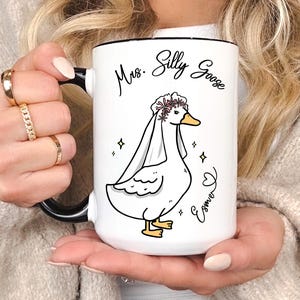 A blonde lady holding her Mrs Silly Goose mug gift that was gifted to her for her engagement party, wedding party or bridal shower. It appears to be the 15oz variant and allows for personalization.