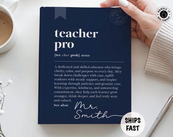 May include: A navy blue notebook with the words "teacher pro" in white. The definition of "teacher pro" is written below. The notebook is held by a person's hand. A white mug and pen are also visible.