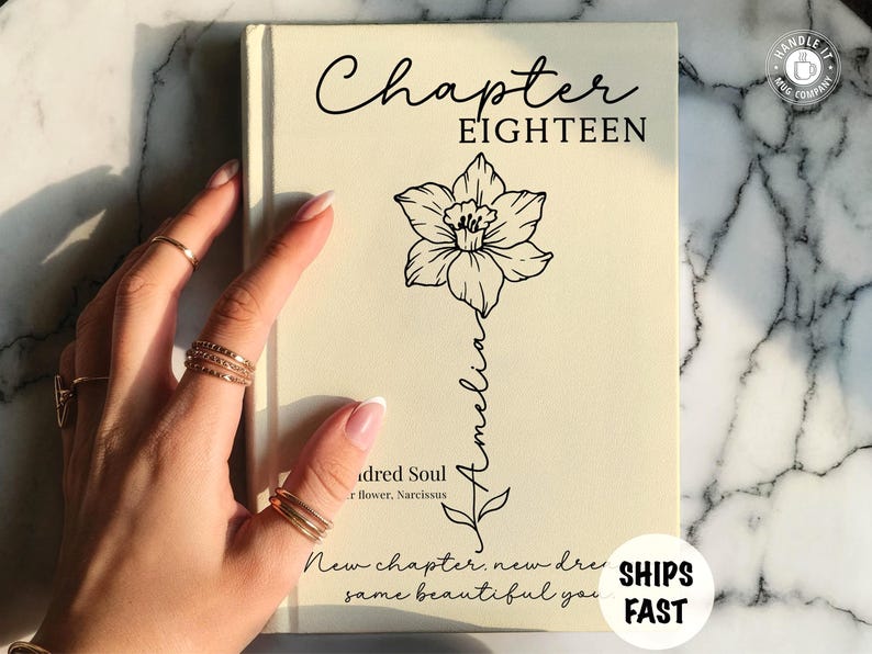May include: A cream-colored book cover featuring the text Chapter EIGHTEEN at the top. A black line drawing of a daffodil flower is centered, with the name Amelia written vertically along the stem. Additional text includes Kindred Soul, flower, Narcissus, and New chapter, new dreams, same beautiful you.