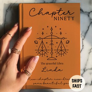 Può includere: Un libro con copertina rigida color arancione bruciato con il testo Chapter NINETY, un'illustrazione a linee nere della bilancia dello zodiaco, la frase The graceful Libra, il nome Linda e il testo New chapter, new dream, same beautiful you.