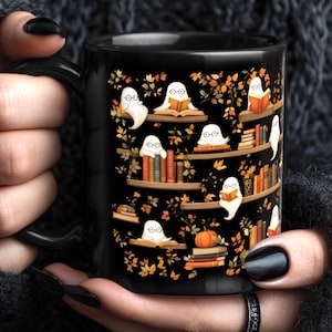 Op de afbeelding: Zwarte keramische mok met een ontwerp van geesten die boeken lezen op boekenplanken, met oranje en gele herfstbladeren en een pompoen. De mok heeft een zwart handvat en de woorden "HANDLE IT MUG COMPANY" in een cirkel.