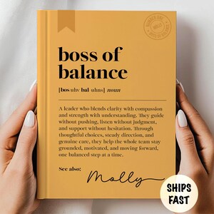May include: Mustard-colored book with the words "boss of balance" in black. The book includes a definition, the name "Molly", and a "Ships Fast" sticker. A mug company logo is in the upper right corner.