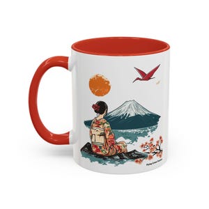 May include: White ceramic mug with a red rim and handle. The mug features a colorful illustration of a woman in a kimono sitting on a rock with Mount Fuji in the background. A red bird flies above the mountain. The text "Flying Crane Factory" is printed on the bottom of the mug.