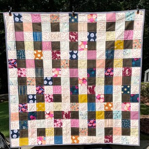 May include: A handmade quilt with a patchwork design. The quilt features a grid pattern of squares in various colors and patterns, including pink, brown, blue, and floral prints. The quilt is bordered with a white fabric with small pink polka dots. The quilt is hanging outside.