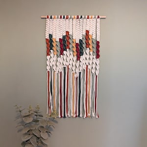 May include: A macrame wall hanging with a wooden dowel. The design features white, red, green, and orange knotted sections. Below, long strands of matching colours hang down. The wall hanging is displayed against a neutral wall.