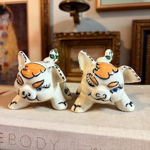 May include: Two ceramic pig figurines. The pigs are white with orange and black markings, blue eyes, and green leaf accents. They are lying down on a book with the word "BODY" visible.