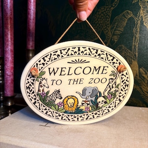 May include: Cream-coloured oval ceramic sign with the text "WELCOME TO THE ZOO" and animal illustrations. The sign has a decorative black border and a jute string for hanging. The sign features a giraffe, zebra, lion, elephant, and monkey.