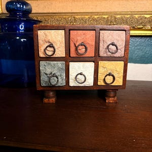 May include: A small, wooden chest of drawers with six square drawers. Each drawer is a different color, including beige, red, gray, white, and gold. The drawers have small, round, black metal pulls. The chest sits on four small, wooden feet.
