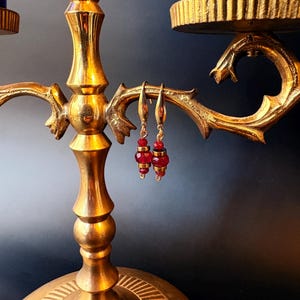 May include: A pair of red and gold beaded earrings hanging from a gold candelabra. The earrings have small red beads and gold accents. The candelabra is ornate with a gold finish and a dark background.