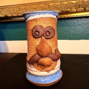 May include: A whimsical, handmade ceramic mug with a face design. The mug features brown, textured clay with applied facial features, including large, round eyes, a nose, and a mustache. The rim and base are painted blue and white.