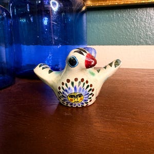 May include: A ceramic bird figurine with outstretched wings. The bird is primarily white with a blue, red, and green beak and accents. The bird's chest features a sun-like design with a yellow face and blue floral details.